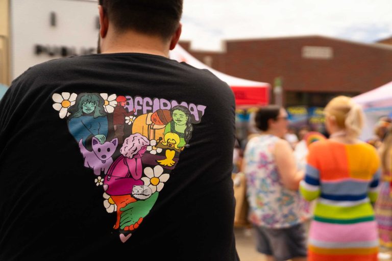 Image of a man at an outdoor event wearing a black t-shirt with a colorful triangle on the back, demonstrating inclusivity.